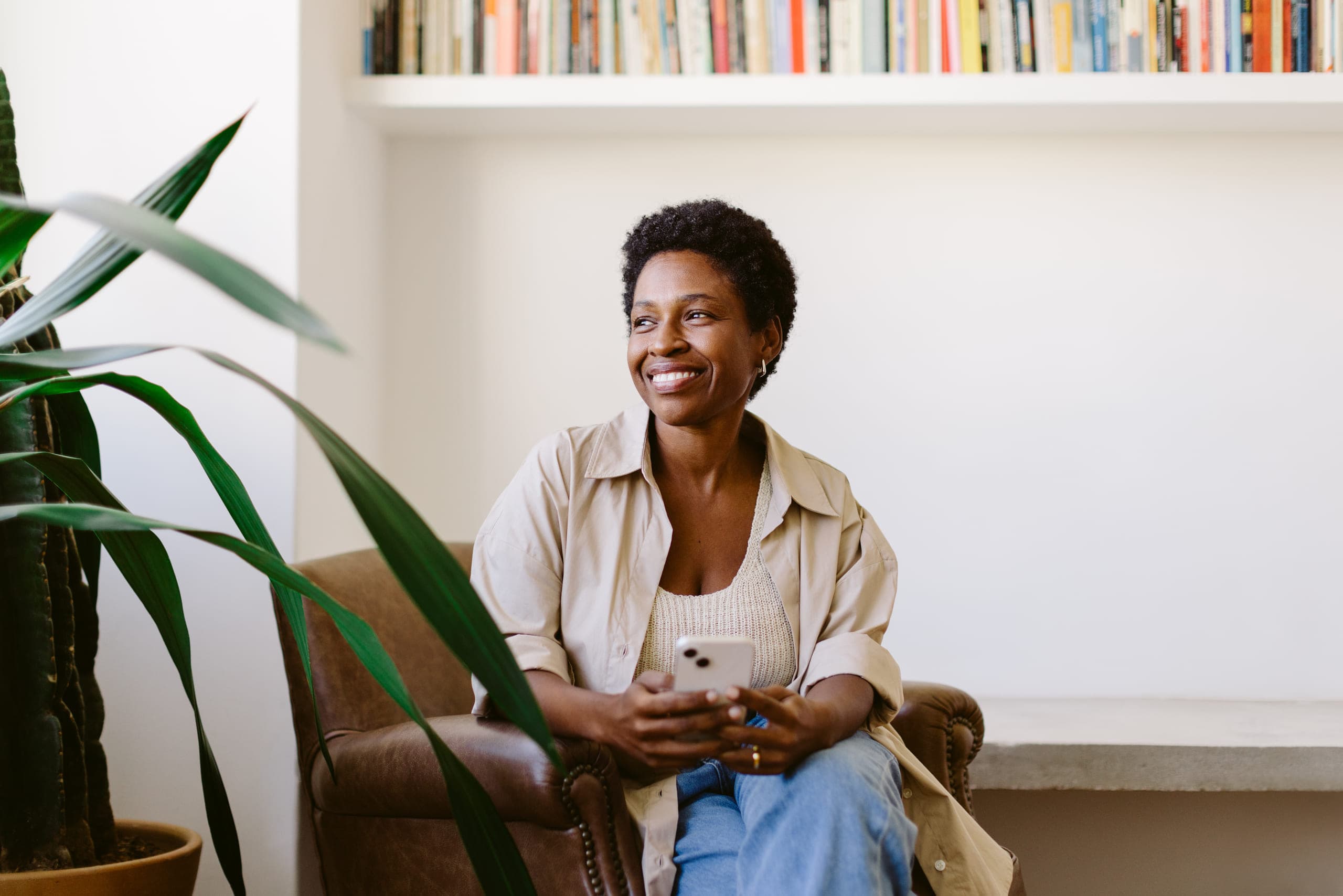 A woman sitting in an armchair at home, holding her phone and smiling as she looks away from the camera.